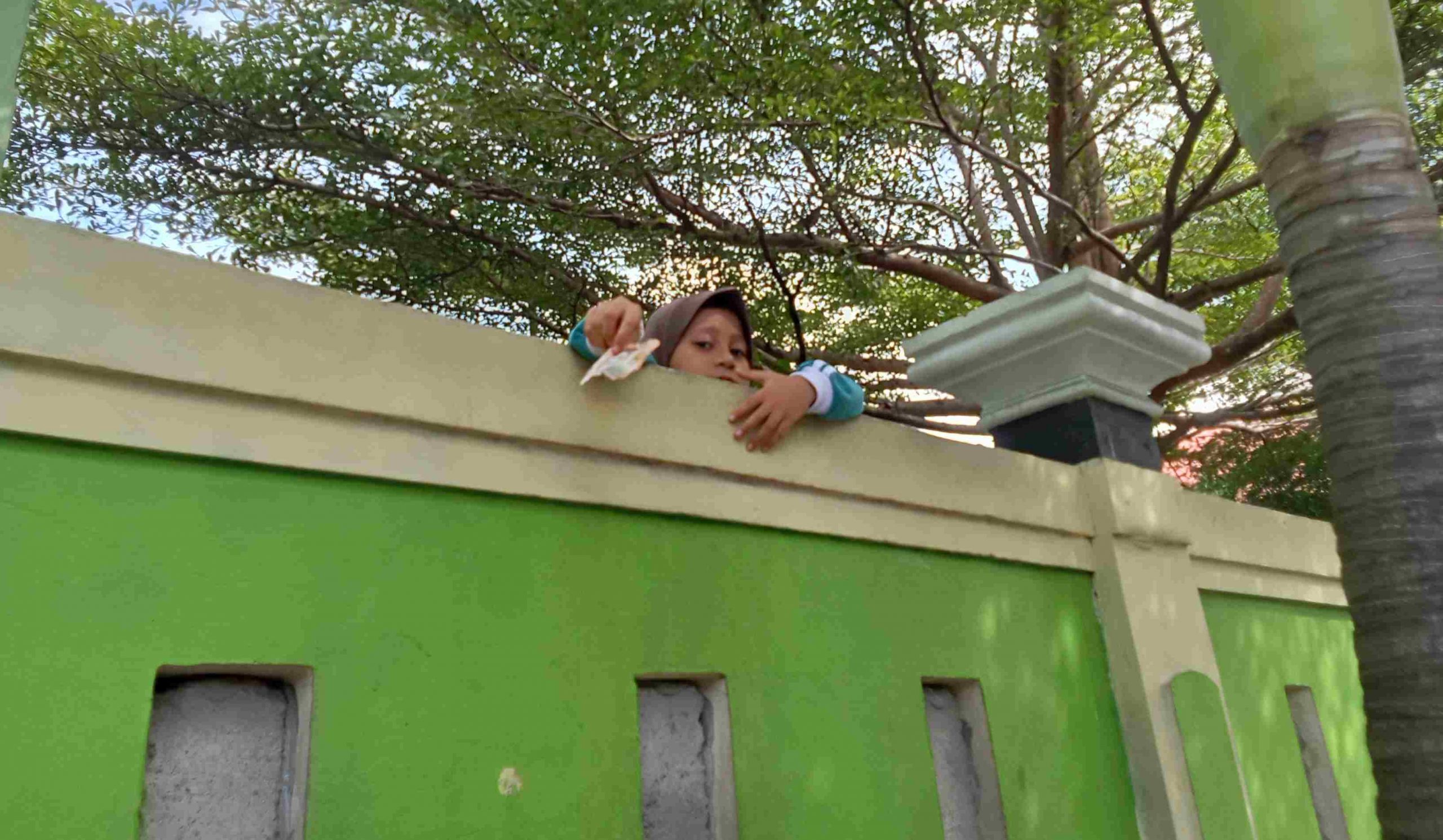 Seorang Siswi terlihat berusaha membeli Jajanan memanjat dan mengulurkan tangan keluar tembok. Terlihat juga lubang Tembok yang sudah di semen.